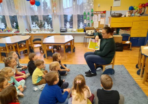 Dzieci siedzą na dywanie, pani bibliotekarka pokazuje ilustracje w książce.